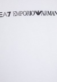 T-shirt en coton blanc arborant un logo "EA7 Emporio Armani" en noir audacieux sur le devant, avec des styles et tailles de police variés.