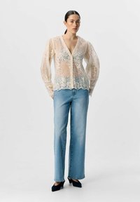 Gennemsigtig bluse af blonde har en dyb V-hals, lange puffærmer og en bølget kant. Kombineret med lyseblå lige jeans og sorte heels.