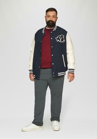 Marineblauwe varsityjack met witte leren mouwen, NYC-logo, grijs sweatshirt, grijze broek en witte sneakers. Gelaagde stoffen en gestreepte accenten.