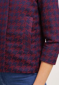 Gros plan d'une manche et d'une poche de veste texturées rouge et bleu marine, portées sur un jean bleu au bras et à la taille d'une personne.