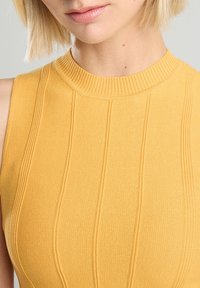 Keltainen, ribattu villapaita korkealla kauluksella, jossa on pystysuoria raitoja ja teksturoitu neulekuvio. Lähikuva kangasdetailista.