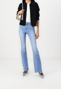 Černá pletená bunda, bílá košile, světlé modré zvonové džíny a metalické stříbrné lodičky. Modelka drží malou stříbrnou kabelku.