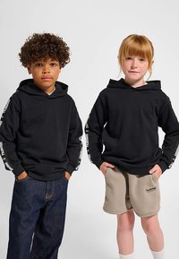 Sorte hættetrøjer med hvidt logo langs ærmerne, båret af et barn i mørke jeans og et andet i beige shorts med hvide sokker.