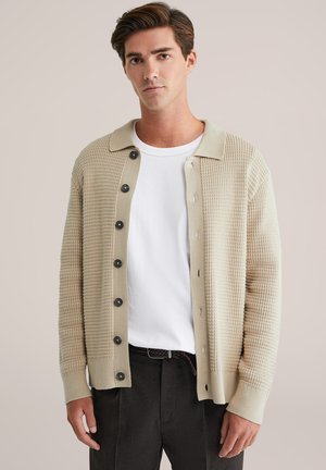 Cárdigan de punto beige con un diseño texturizado, con cierre de botones, puños acanalados y un cuello claro en contraste sobre una camisa blanca.