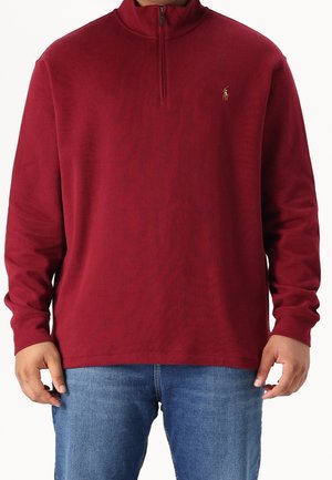 Mand iført en langærmet, vinrød quarter-zip pullover med et lille broderet logo på brystet og blå jeans.