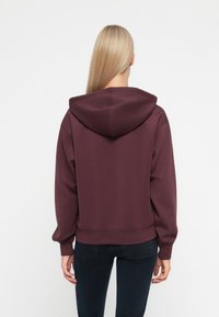 Bordowy pulower z kapturem, wykonany z miękkiego materiału, z przestronnym kapturem, ściągaczami przy rękawach i skróconym krojem. Brak widocznych grafik lub wzorów.