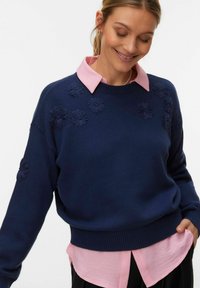Femme souriante regardant vers le bas, portant un pull bleu marine avec des détails floraux texturés, sur une chemise rose à col, et un pantalon noir.