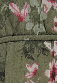 Blommigt tyg med rosa och mörka blommor på en olivgrön bakgrund, med en texturerad yta och en rynkad sömdetalj.