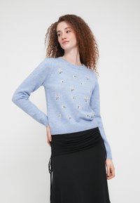 Ανοιχτό μπλε πουλόβερ με στρογγυλή λαιμόκοψη, με κεντήματα λουλουδιών σε χρυσό και λευκό. Συνδυάζεται με μαύρη φούστα με ρεγκλάν.