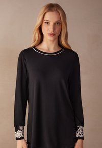 Schwarzes Langarmshirt mit rundem Halsausschnitt und silbernem Akzent. Die Spitzenverzierungen an den Bündchen verleihen Struktur und Kontrast. Glattes Material.