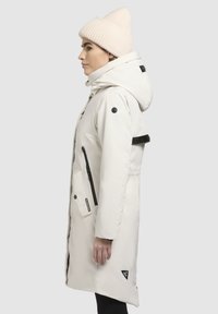 Witte waterdichte jas met capuchon, zwarte accenten en ritszakken. Gecombineerd met een geribbelde crèmekleurige beanie, slanke vorm en zachte textuur.