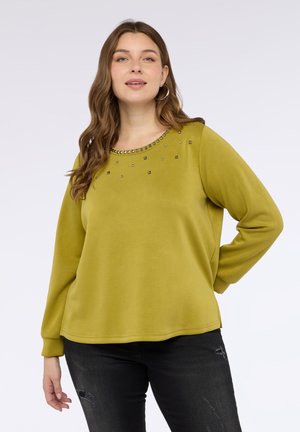 Sudadera de color verde lima con un cuello redondo adornado con tachuelas metálicas, mangas largas y un corte holgado, combinada con jeans negros desgastados.