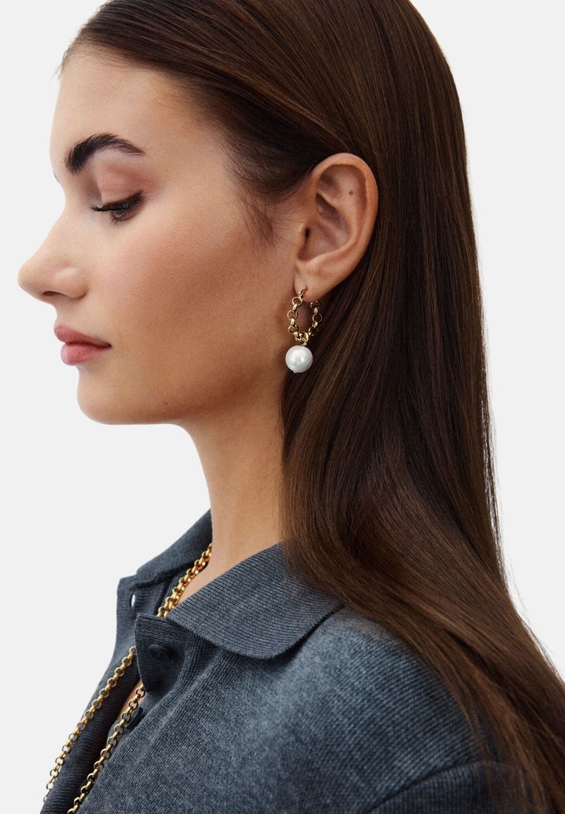 Pendientes de aro dorados con enlaces entrelazados, adornados con una gran perla blanca. El modelo lleva un suéter de cuello en gris oscuro.