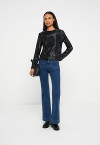 Femeie stând în fața unui fundal alb, purtând un top negru texturat cu mânecă lungă, blugi albaștri, pantofi loafer negri și ținând o geantă mică neagră.