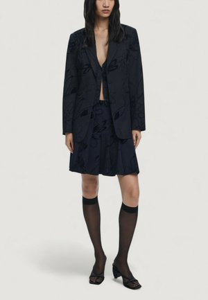 Black floral-printed blazer with a deep V-neck, paired with a matching skirt. Model wears black knee-high stockings and open-toe heels.