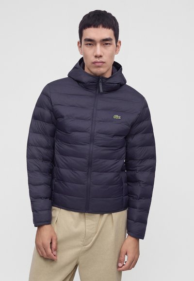 Jeune homme portant une veste matelassée à capuche bleu marine foncé avec fermeture éclair à l'avant et un pantalon beige, debout devant un fond clair uni.