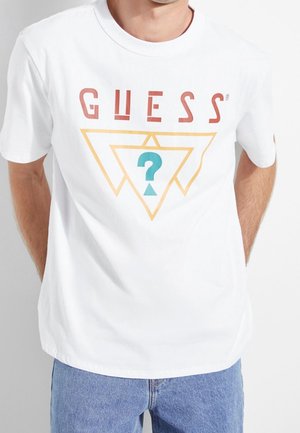 Hombre con una camiseta blanca con el texto "GUESS" sobre un triángulo dorado superpuesto y un signo de interrogación azul dentro del triángulo.