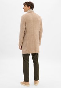 Beige Corduroy-Mantel mit Reverskragen, langen Ärmeln und geradem Schnitt. Kombiniert mit dunkelgrünen Hosen und hellbraunen Schuhen.