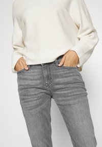 Jeans de mezclilla gris con un corteSlim y una textura sutil. Combinados con una parte superior de manga larga crema. Dispone de bolsillos laterales y un cierre frontal con cremallera.