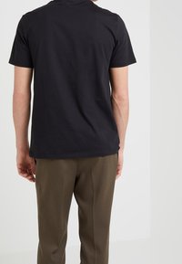 Man som bär en svart t-shirt med kort ärm och olivgröna byxor, står vänd mot en plain ljusgrå vägg.