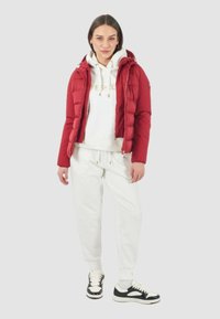 Giacca a vento rossa con baffle verticali, felpa crema con testo dorato, pantaloni sportivi bianchi e sneakers nero e bianche. Outfit casual e a strati.
