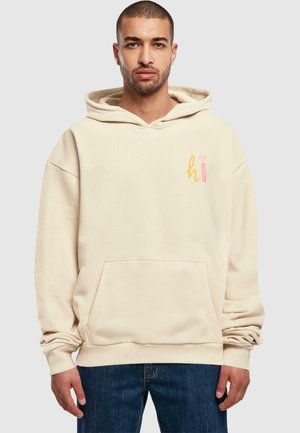 Beige hættetrøje med en forlommeklud, snor i hætten og farverig tekst "hi" trykt på brystet. Båret med mørkeblå jeans.