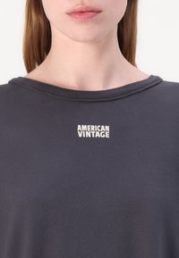 Mørk grå bomull T-skjorte med vid utringning og en liten hvit "AMERICAN VINTAGE" logo på brystet. Glatt tekstur, avslappet passform.