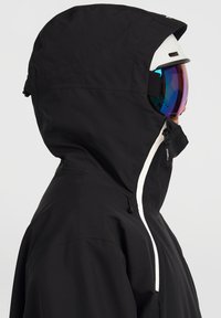 Giacca da sci nera impermeabile con cappuccio, dettagli visibili in bianco sulla cerniera e una tasca laterale. Indossata con un casco e occhiali da sci a specchio.