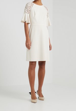 Femme portant une robe crème longueur genoux avec des manches courtes en dentelle et des poignets évasés, associée à des talons beiges avec des nœuds noirs.