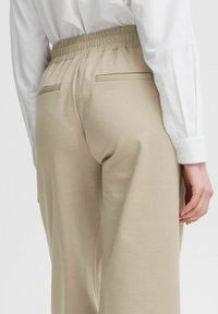 Beige vida byxor med elastisk midja, sidofickor och en slät yta, parat med en vit långärmad skjorta med knappar.