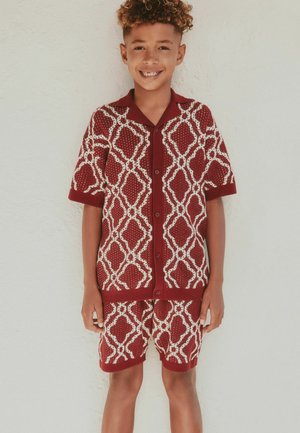 Băiat zâmbitor cu păr creț, purtând o cămașă cu mâneci scurte și pantaloni scurți asortați, roșii cu alb, stând în fața unui perete deschis la culoare.