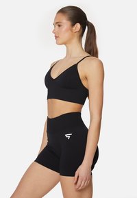 Črn športni modrček s tankimi naramnicami in rahlim v-izrezom, v kombinaciji z visokim črnim šortsem, ki na strani nosi belo logo.
