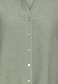 Blouse vert clair avec un décolleté en V et six boutons blancs, présentant un tissu texturé et de subtils détails de couture le long de la patte de boutonnage.