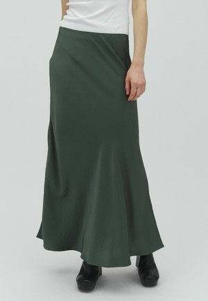 Kobieta ubrana w ciemnozieloną długą spódnicę, biały top bez rękawów oraz czarne botki na platformie, stojąca na jednolitym jasnym tle.