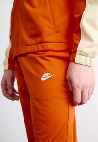 Nike φόρμα με φωτεινό πορτοκαλί ύφασμα με απαλό υφή, αντίθετη κρέμα απόχρωση στην μανσέτα και λευκή λεπτομέρεια λογότυπου.
