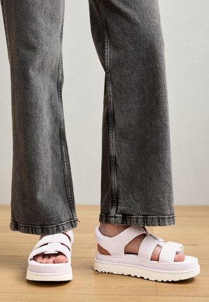 Lys rosa sandaler med flere stropper, teksturert materiale og en tykk hvit såle. Kledd med grå vide bukser.