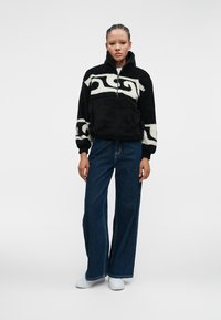 Sort fleece pullover med hvidt abstrakt mønster, halvlukket forsygning og lange, let puffede ærmer, parret med vide denim bukser.