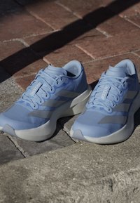 Para jasnoniebieskich butów do biegania Adidas Adizero z białymi podeszwami umieszczonych na kamienno-ceglanym chodniku w świetle słonecznym.