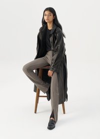 Black leather trench coat with buttons and belt detail, paired with light brown fitted pants and black loafers, seated on a wooden stool.