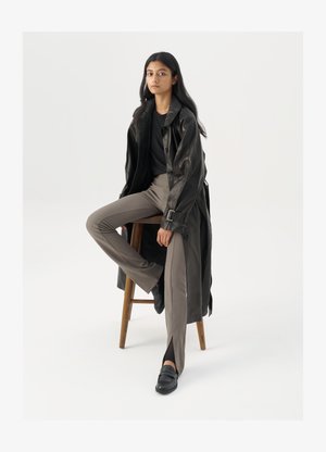 Must musta trench-jakk mustriga nuppudega ja vöödetailiga, koos heledatest pruunidest sobivaist pükstest ja mustadest loafers'itest, istudes puidust toolil.