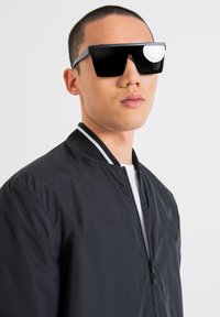 Occhiali da sole neri oversize con lenti piatte, montatura spessa e dettaglio riflettente. Indossati con un giubbotto bomber nero con un colletto a contrasto.
