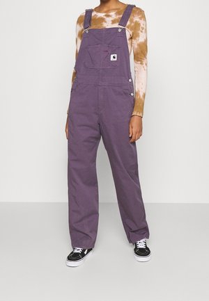 Henkilö, joka käyttää löysiä violetteja haalareita ruskean ja valkoisen tie-dye -pitkähihainen paidan päällä sekä mustavalkoisia lenkkareita, seisoo yksinkertaisen taustan edessä.