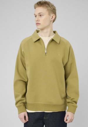 Jeune homme aux cheveux blonds courts portant un sweat-shirt à col zippé vert olive et un jean foncé, regardant vers la gauche sur un fond gris uni.