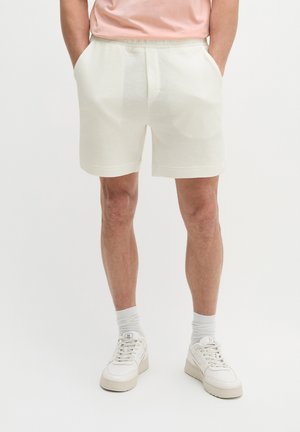 Homme portant un short blanc texturé, un t-shirt rose clair, des chaussettes blanches et des baskets blanches, debout les mains dans les poches.