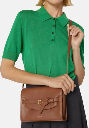 Zelené polo tričko s krátkými rukávy, černými knoflíky a hnědou koženou kabelkou přes rameno s detailním zapínáním na otočný prvek a zlatými kovovými doplňky.