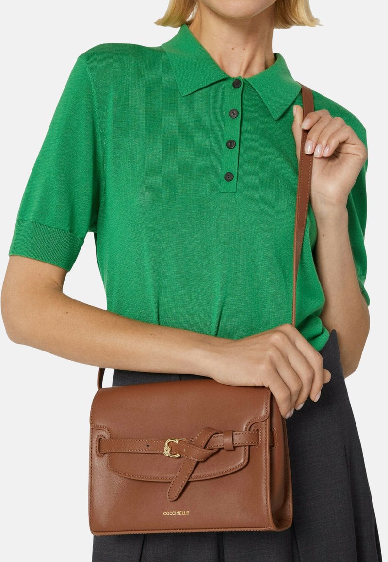Zelené polo tričko s krátkými rukávy, černými knoflíky a hnědou koženou kabelkou přes rameno s detailním zapínáním na otočný prvek a zlatými kovovými doplňky.