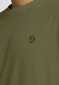 T-shirt a maniche corte color verde oliva realizzata in materiale morbido, con un piccolo logo esagonale nero nella zona del petto.