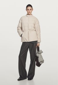 Donna in giacca trapuntata crema, pantaloni larghi scuri, scarpe nere, che tiene una borsa marrone con una sciarpa a scacchi grigia annodata al manico.