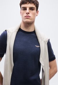 Νεαρός άνδρας που φοράει μπλε σκούρο πλεκτό μπλουζάκι με μπεζ αμάνικο πουλόβερ ριγμένο στους ώμους, στέκεται μπροστά σε απλό φωτεινό φόντο.