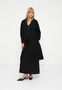 Mulher com cabelo liso loiro, vestindo um longo casaco preto, um vestido preto e sapatos de salto baixo pretos, em pé contra um fundo branco.